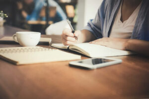 Person writing in a journal with a pen, teacup and phone visible